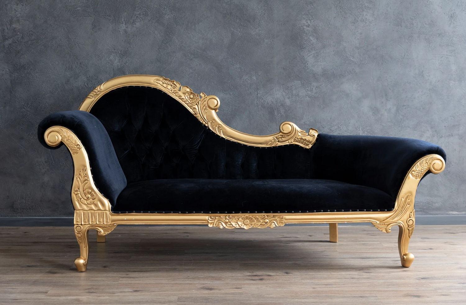 Luxurious black velvet chaise lounge with ornate gold trim against grey wall.