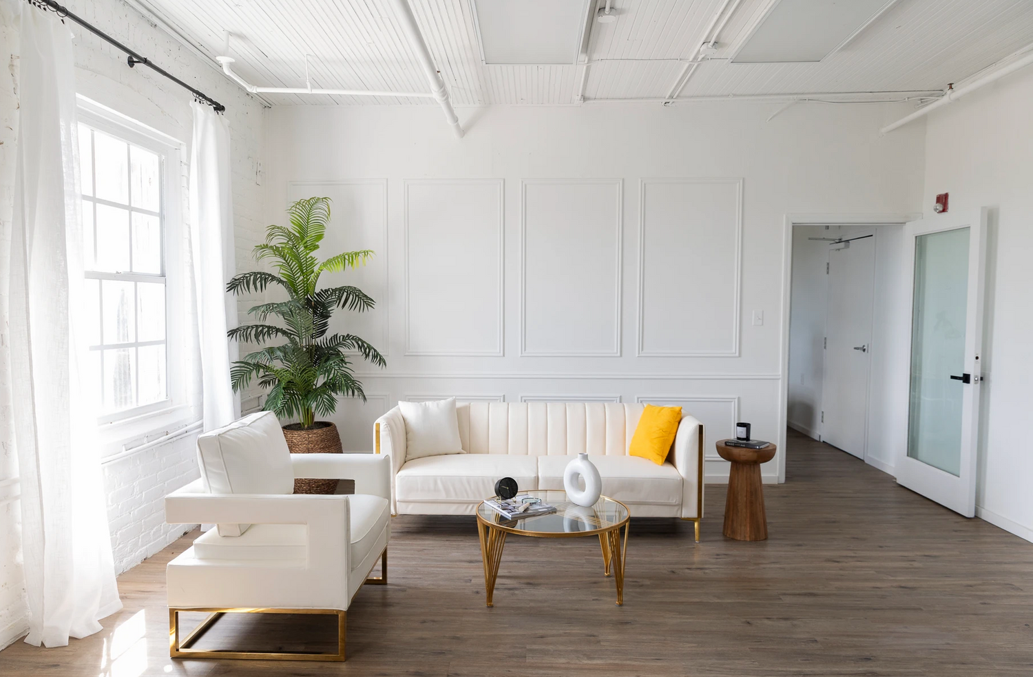 Modern minimalist white living room with sofa, ottoman, wooden accents and potted plant.
