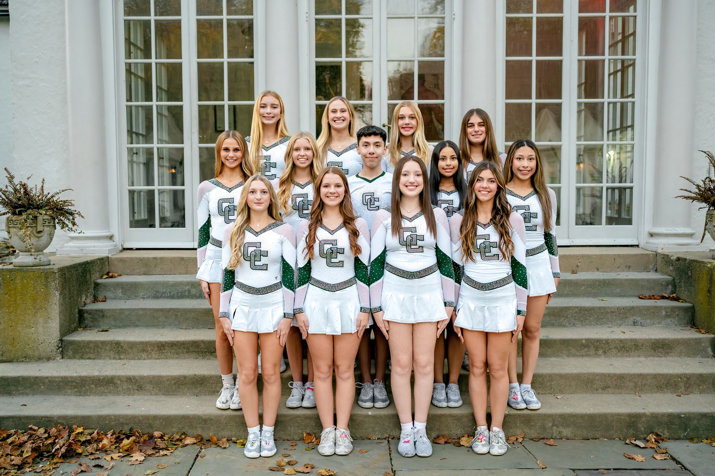 A cheerleading squad in white and green uniforms poses together on stone steps in front of a building.