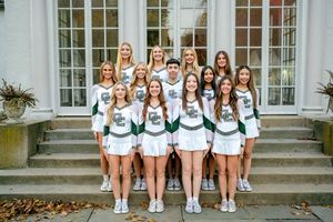 A cheerleading squad in white and green uniforms poses together on stone steps in front of a building.