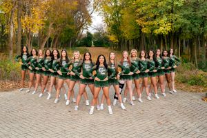 A cheerleading team poses in formation wearing green uniforms during an autumn photoshoot in a wooded setting.
