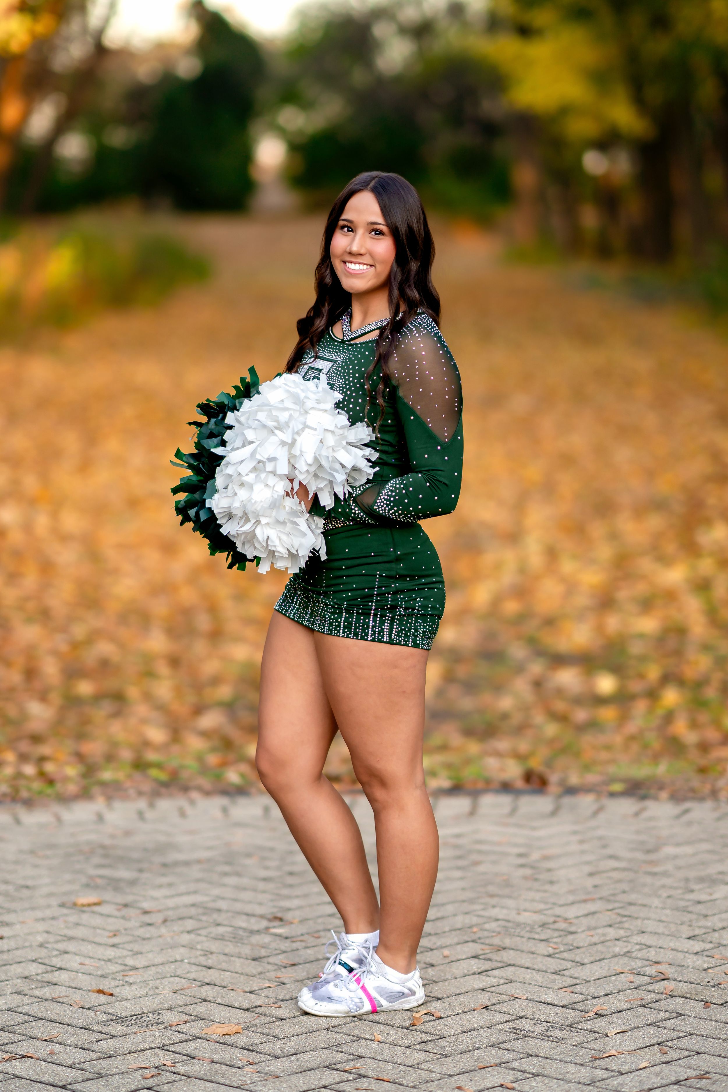 A cheerleader poses in a sparkly green uniform and white sneakers among fallen autumn leaves on a brick path.