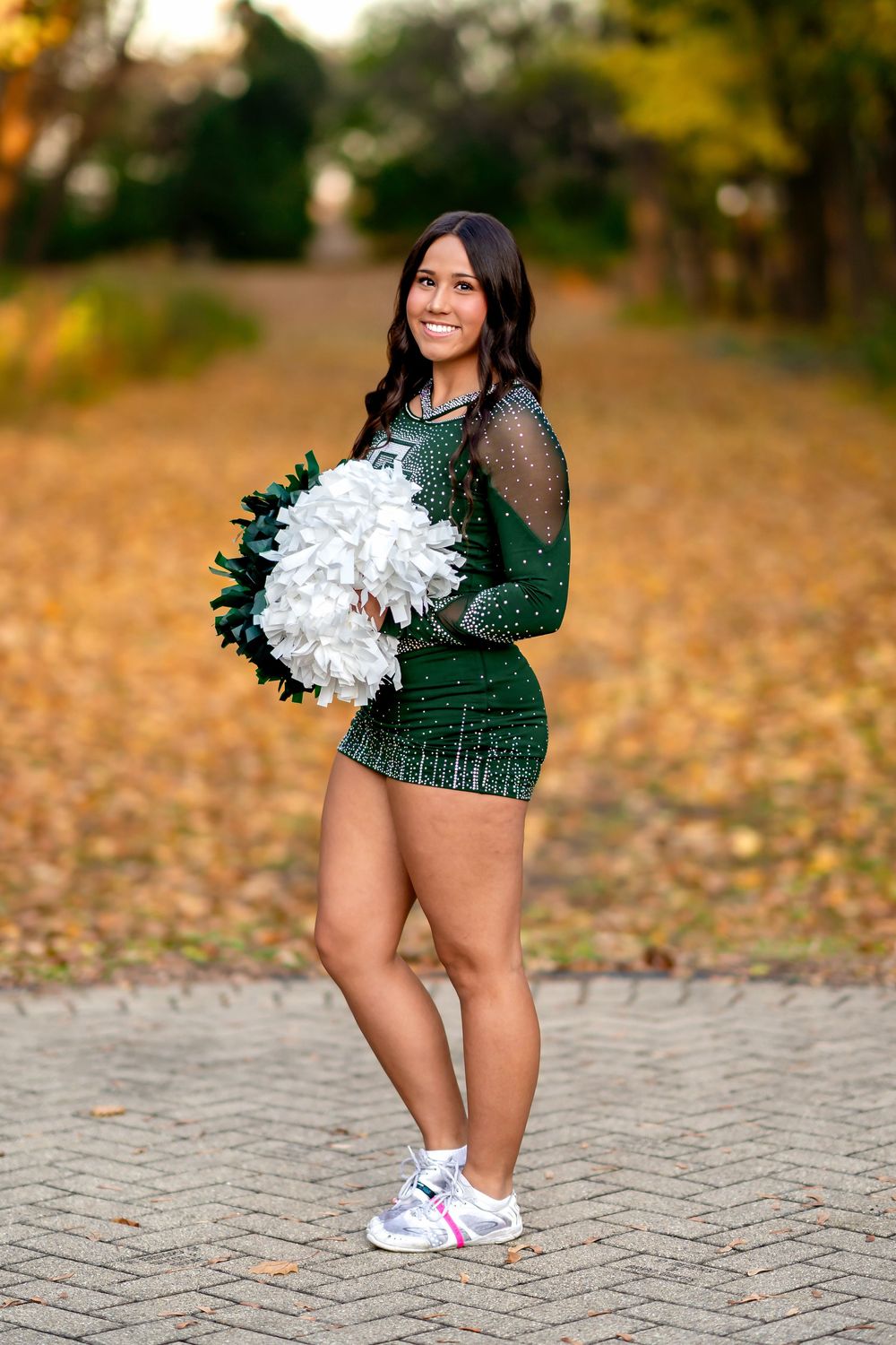 A cheerleader poses in a sparkly green uniform and white sneakers among fallen autumn leaves on a brick path.