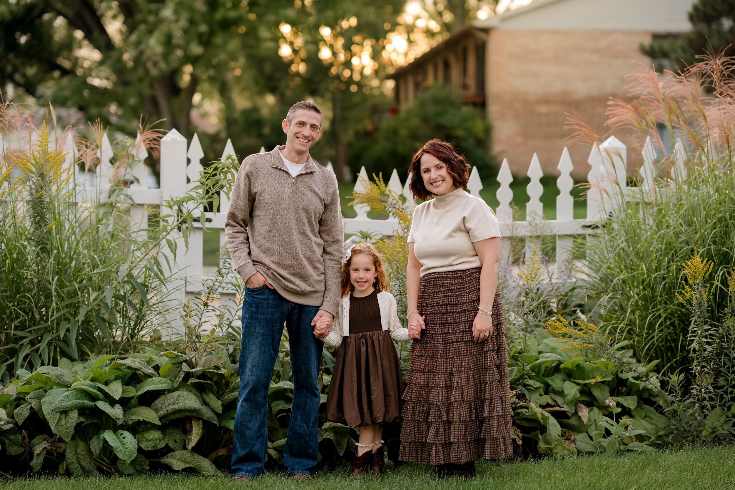 Family of three posing in lush garden by white picket fence at golden hour, parents holding young daughter's hands.