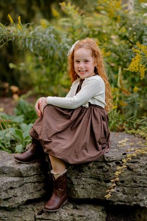 A smiling redhead girl in a brown skirt and cowboy boots sits on a stone wall surrounded by lush green garden foliage.