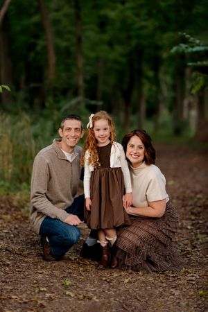 A smiling family of three poses on a wooded trail, with parents kneeling beside their curly-haired daughter in autumn attire.