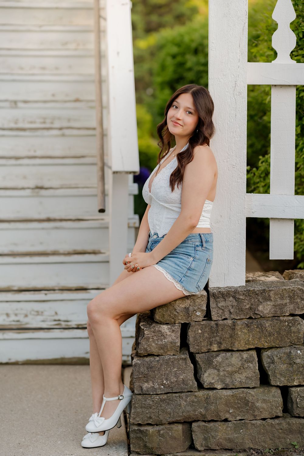 Young woman in denim shorts and white top sits on stone wall by white fence outdoors.