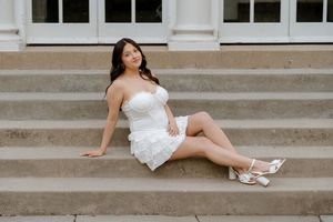 Young woman in white strapless mini dress and heels sitting gracefully on outdoor stone steps.