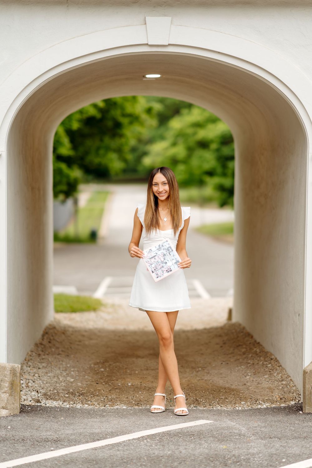 A person in a white summer dress stands in a tunnel-like archway surrounded by greenery on a sunny day.