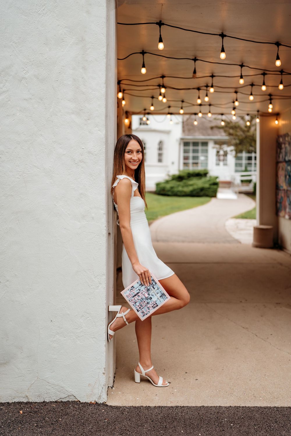 Woman in white dress stands under string lights in covered walkway holding sparkly clutch purse and wearing heels.