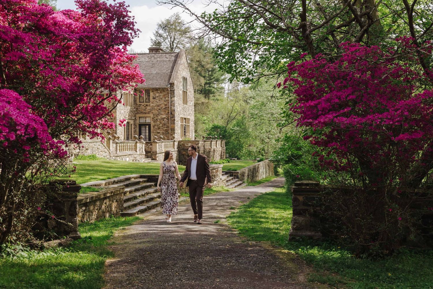Couple strolls along a wooded path in Ridley Creek State Park with pink azaleas and a mansion behind them.
