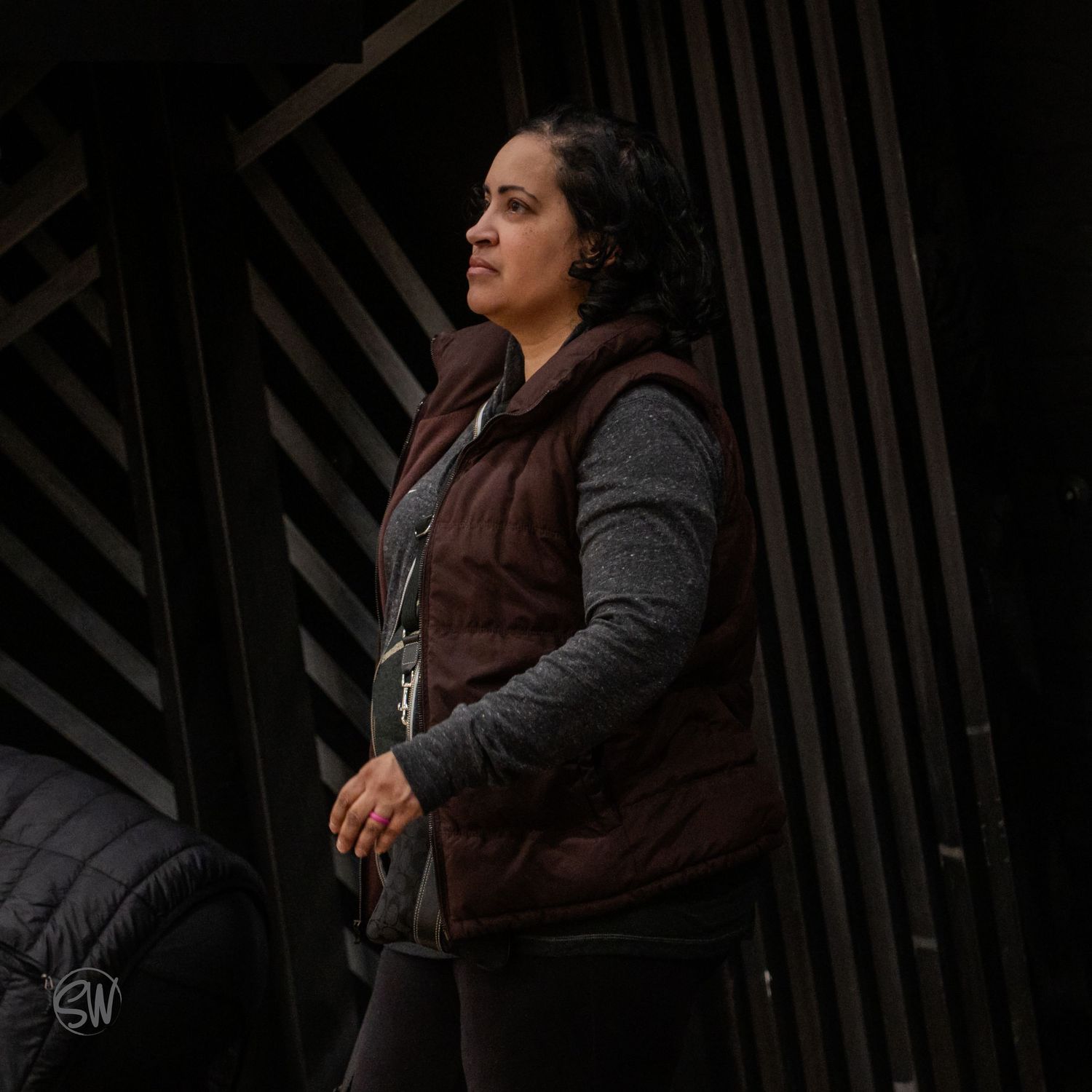 Person wearing a brown vest and gray sweater stands against dark geometric architecture.