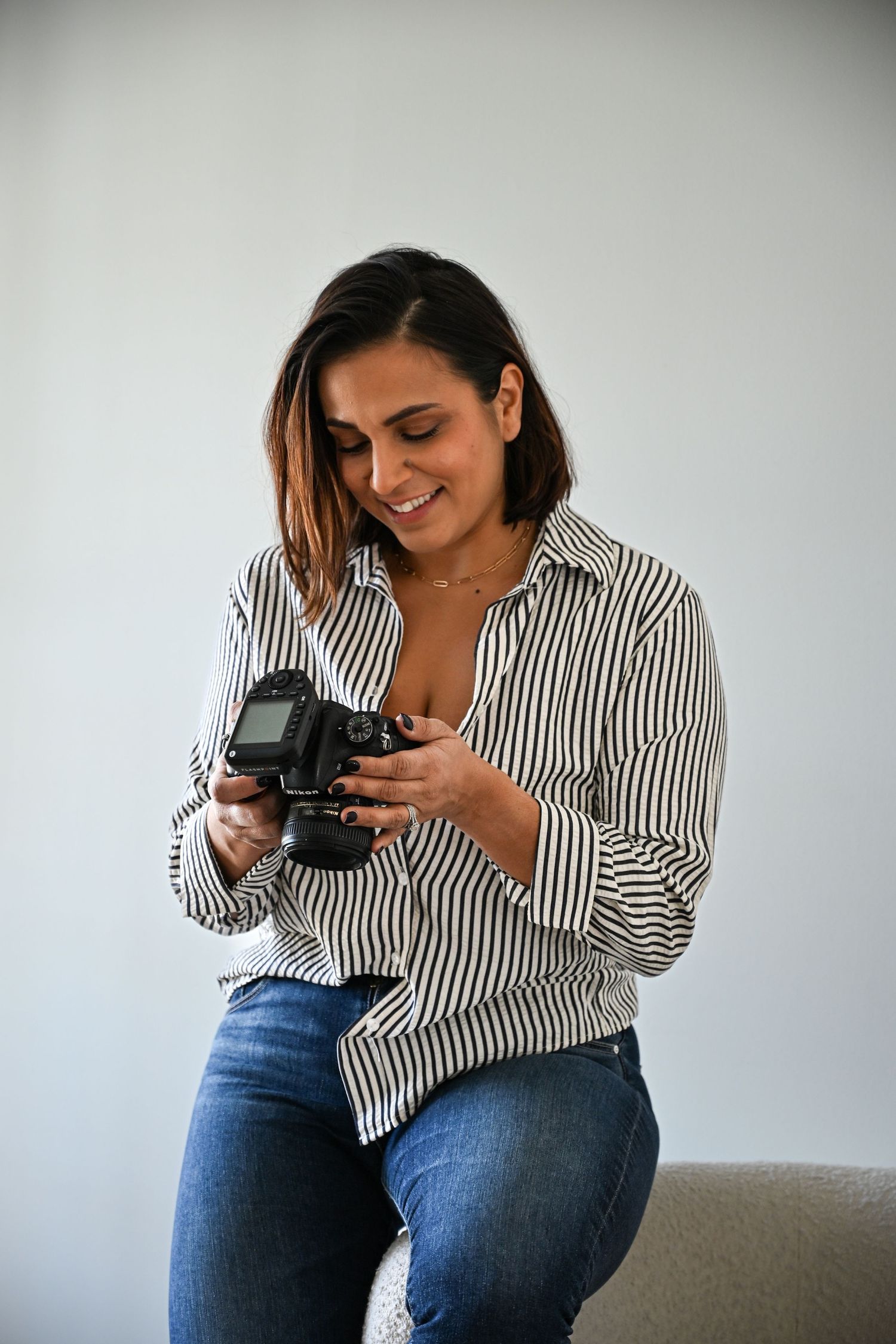 Someone wearing a striped shirt and jeans sits casually while looking down at a digital camera with a smile.