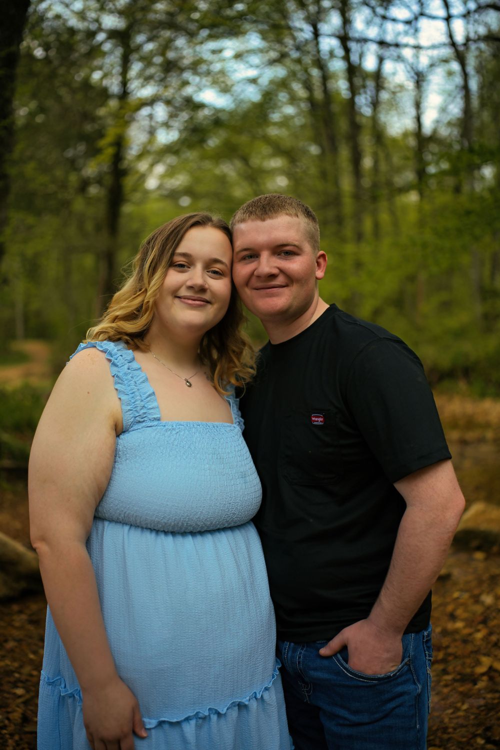 Couple poses together in wooded setting with autumn leaves on ground.