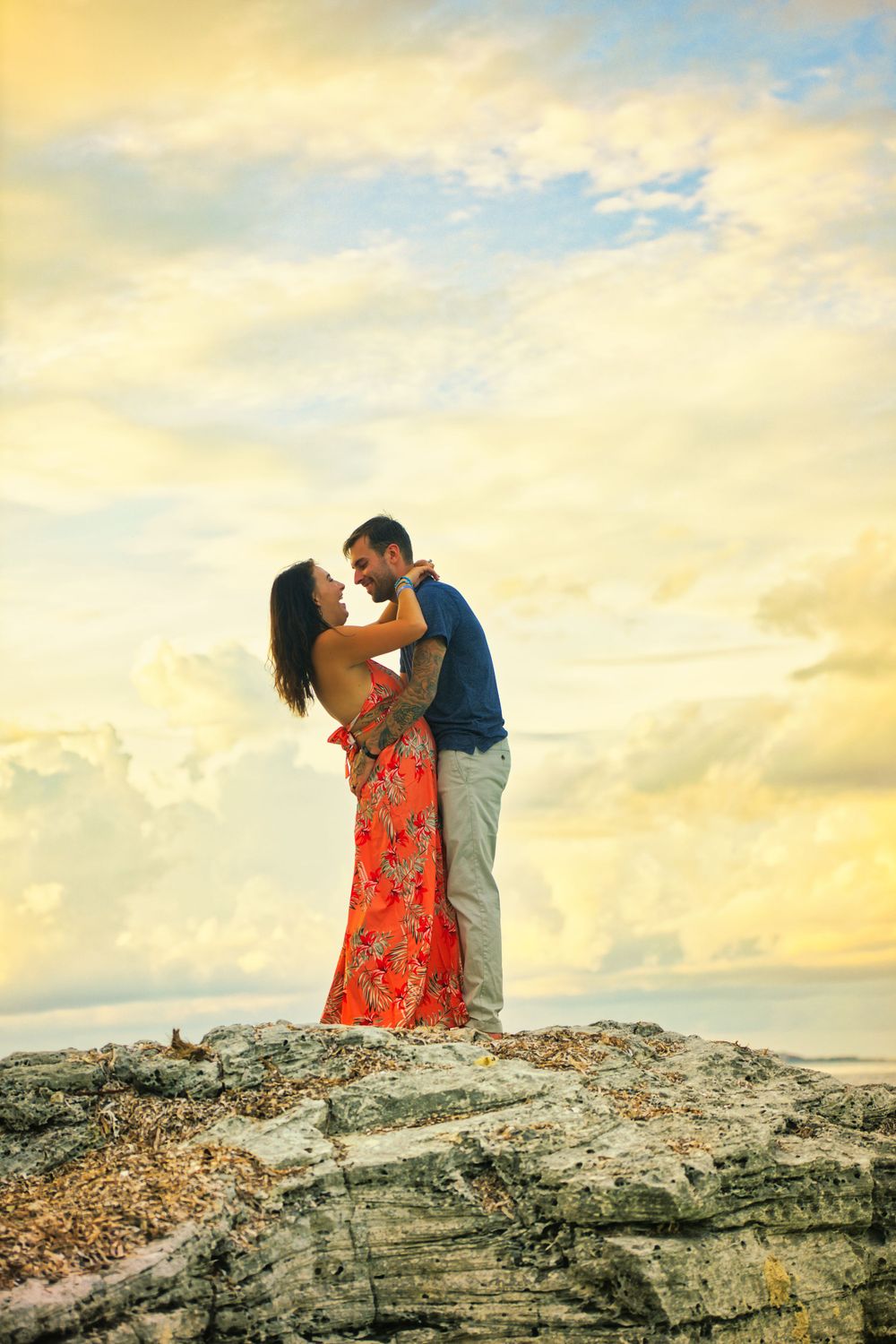 Romantic couple embraces on rocky cliff overlooking ocean at sunset.