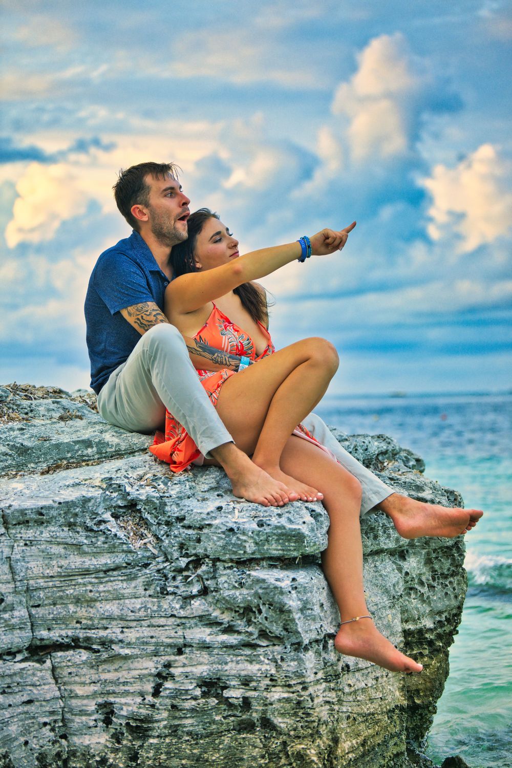 Couple sits on coastal cliff pointing at horizon with dramatic clouds and ocean backdrop.