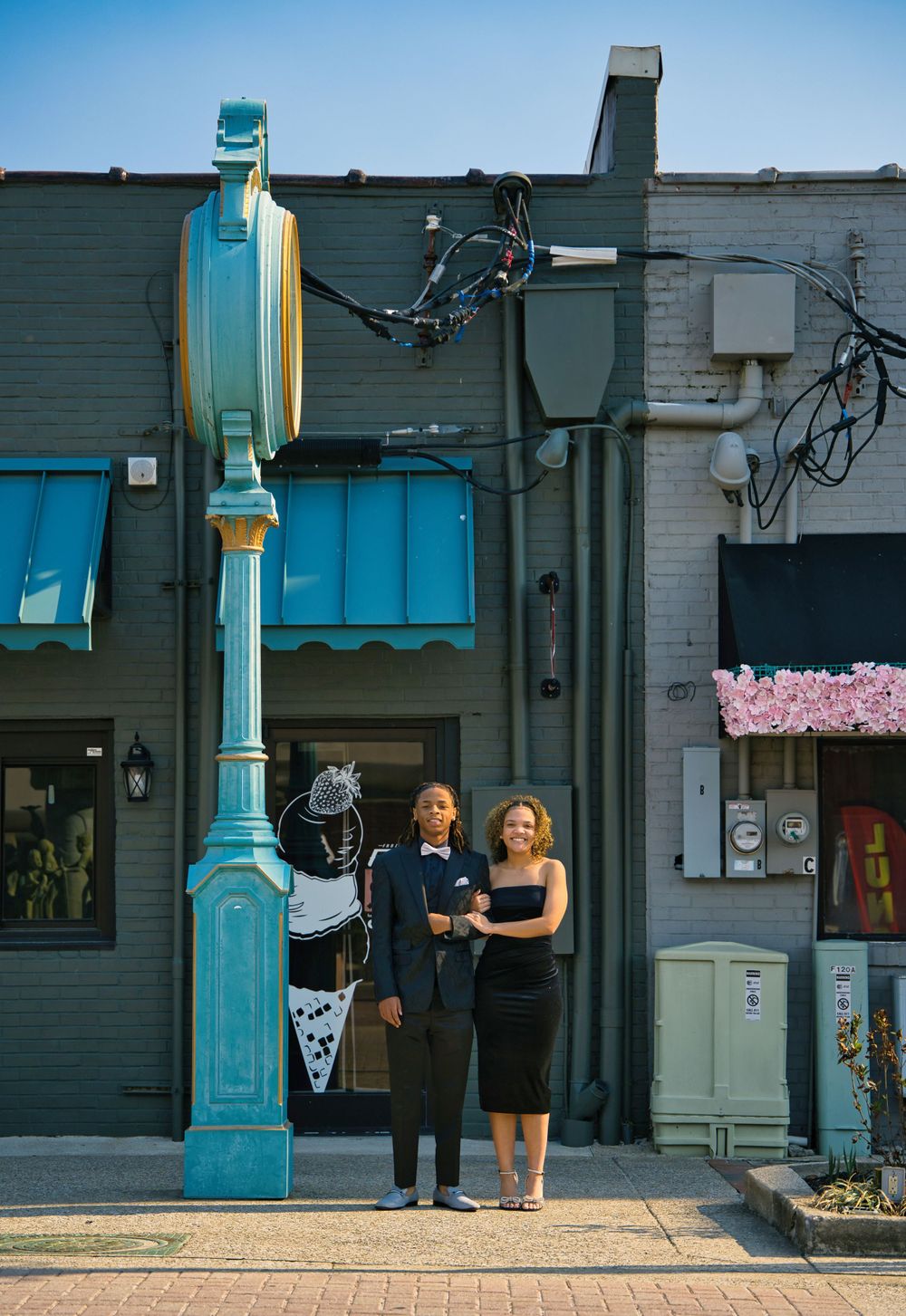 A teal painted decorative lamp post stands in front of a brick building with awnings and storefronts on a paved sidewalk.
