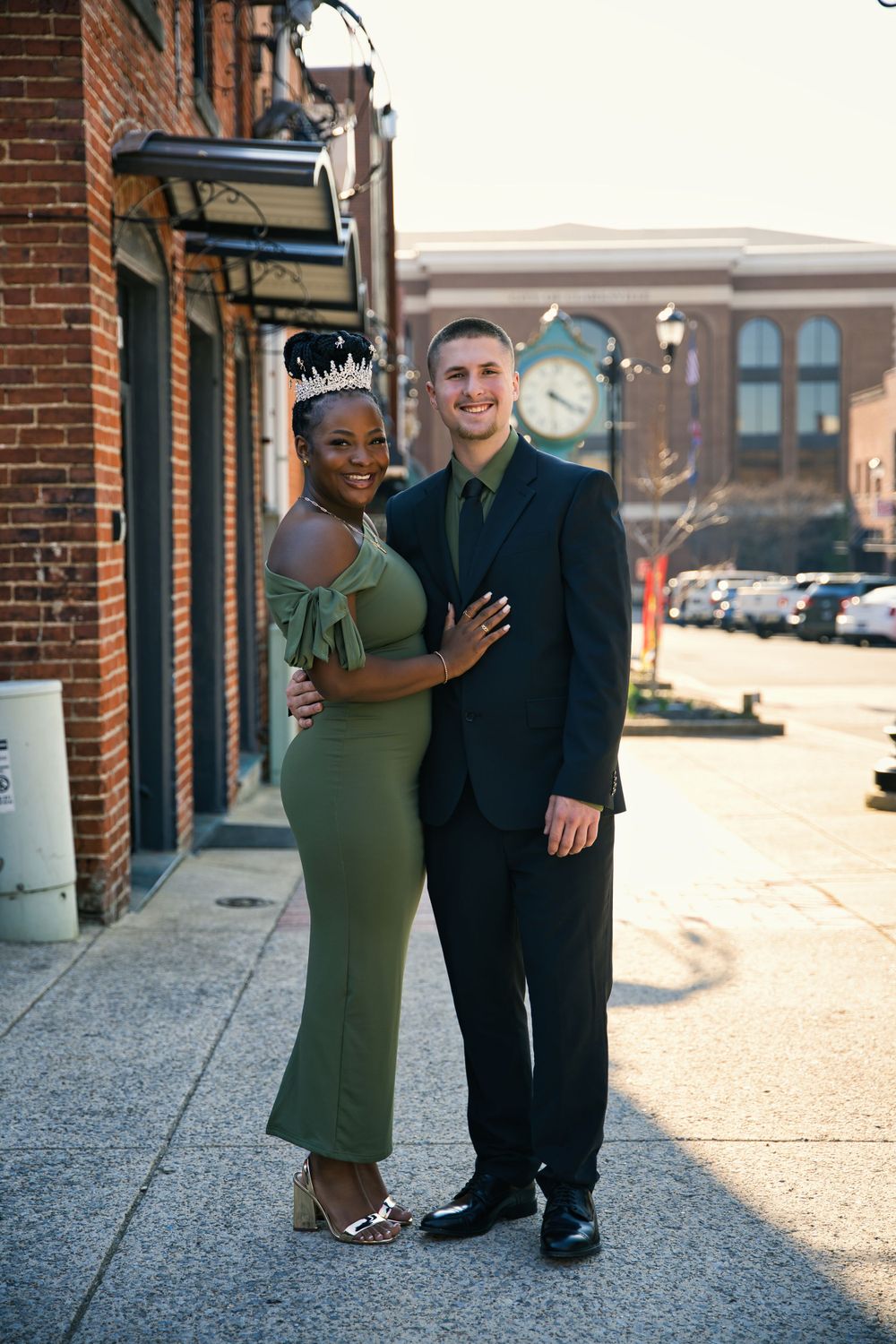 A couple in formal attire poses on a sidewalk outside a brick building on a sunny afternoon.