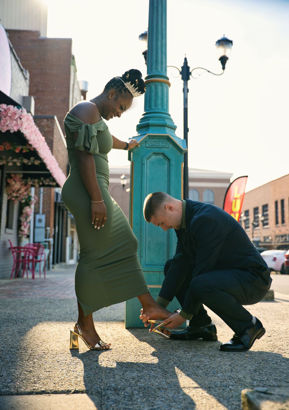A couple shares a romantic moment by a turquoise lamppost on a city street as the sun sets behind buildings.