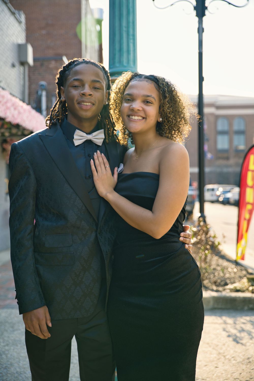 A couple in formal attire poses together outside, with the person in a black tuxedo and bowtie and their date in a black dress.