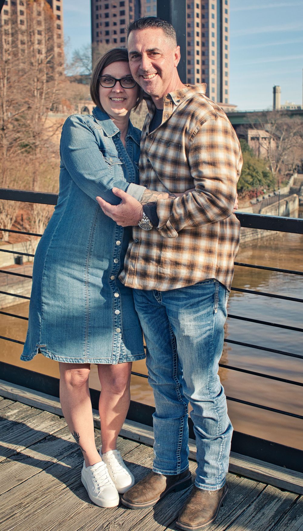 Couple in plaid and denim embracing on wooden deck with urban buildings in background.
