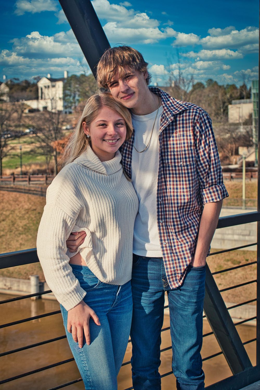 Young couple in casual attire poses together on outdoor deck with cityscape in background.