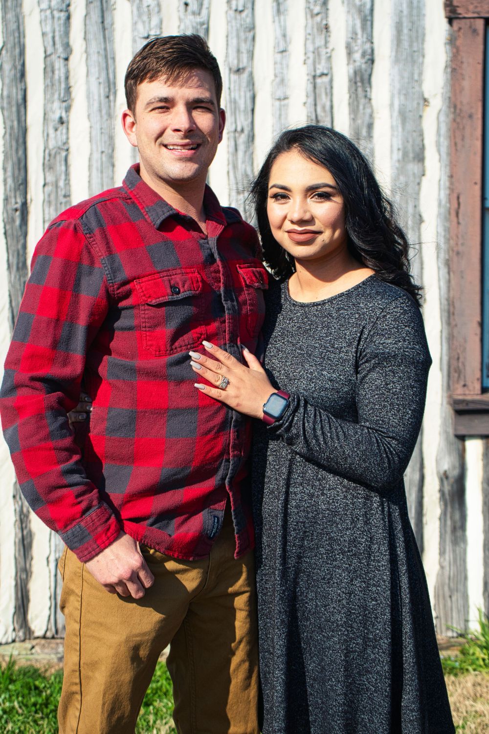 Engaged couple poses together against rustic wall with person in red plaid shirt and gray dress showing engagement ring.