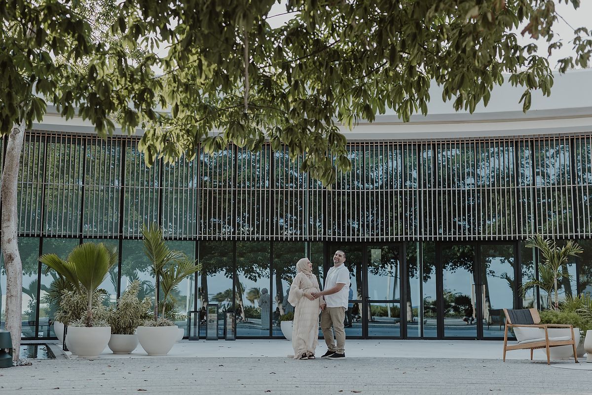 Muslim couple in traditional attire walking outside a modern glass building surrounded by tropical trees.