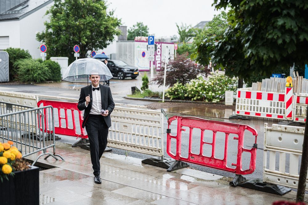 A person in formal attire walks through a rainy street holding an umbrella near red construction barriers.
