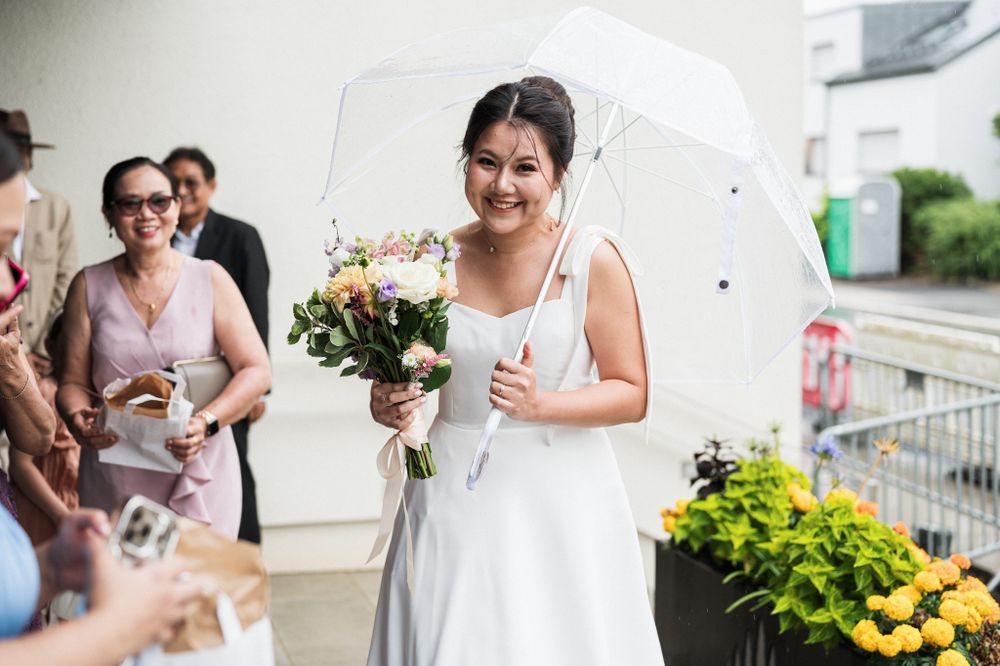 A bride holding a bouquet smiles under an umbrella outside during the wedding celebrations.