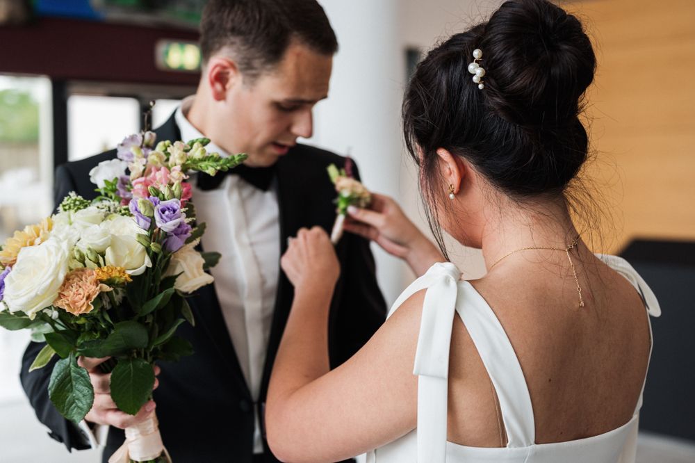 Couple at wedding pinning boutonnieres and holding floral bouquet during intimate wedding preparation moment.