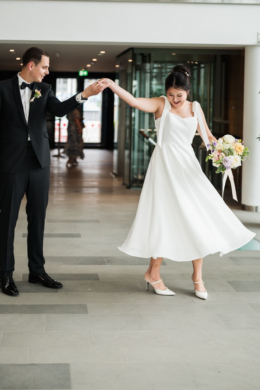 Couple in wedding attire dancing and twirling in modern building hallway with floral bouquet.