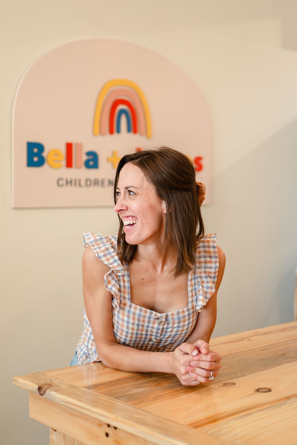 Woman in gingham dress leaning on wooden table smiling in front of colorful rainbow wall art sign.