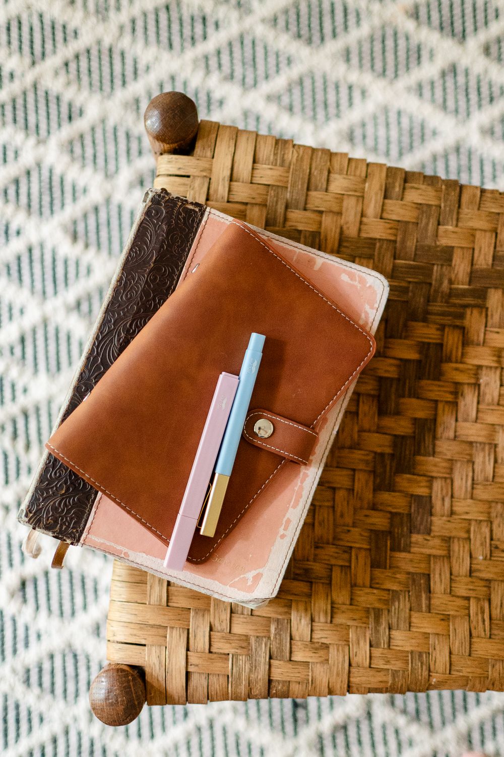 Brown leather wallet with pen on woven bamboo tray against geometric pattern rug.