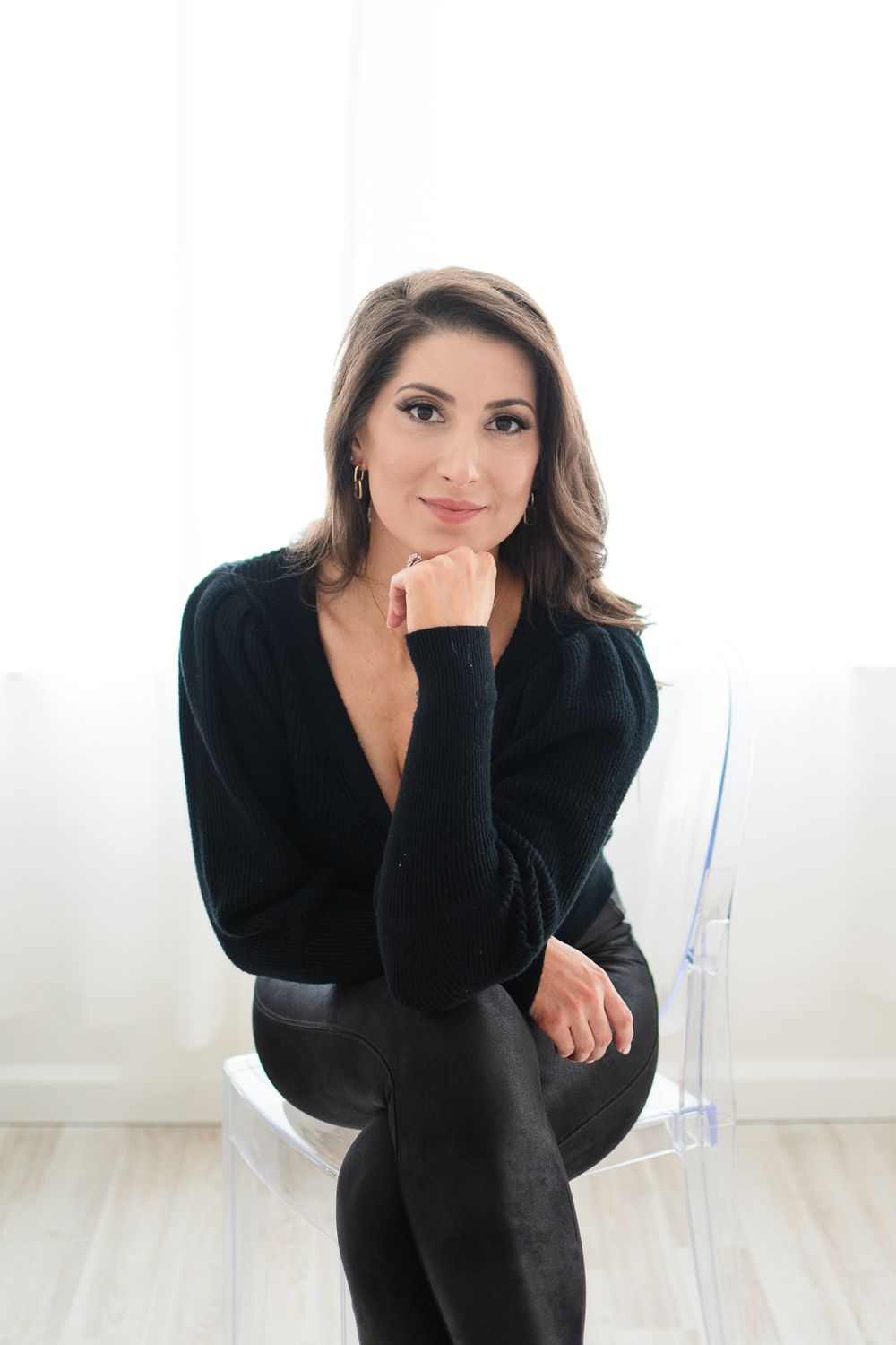 Professional portrait in minimalist setting with clear chair against white wall.