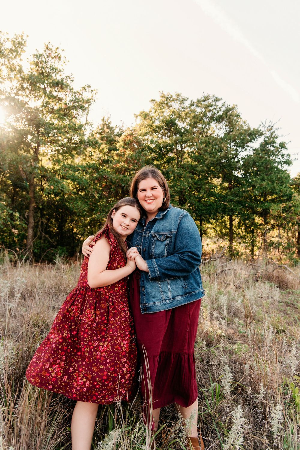Two people embrace while standing in a field with trees in the background at sunset.