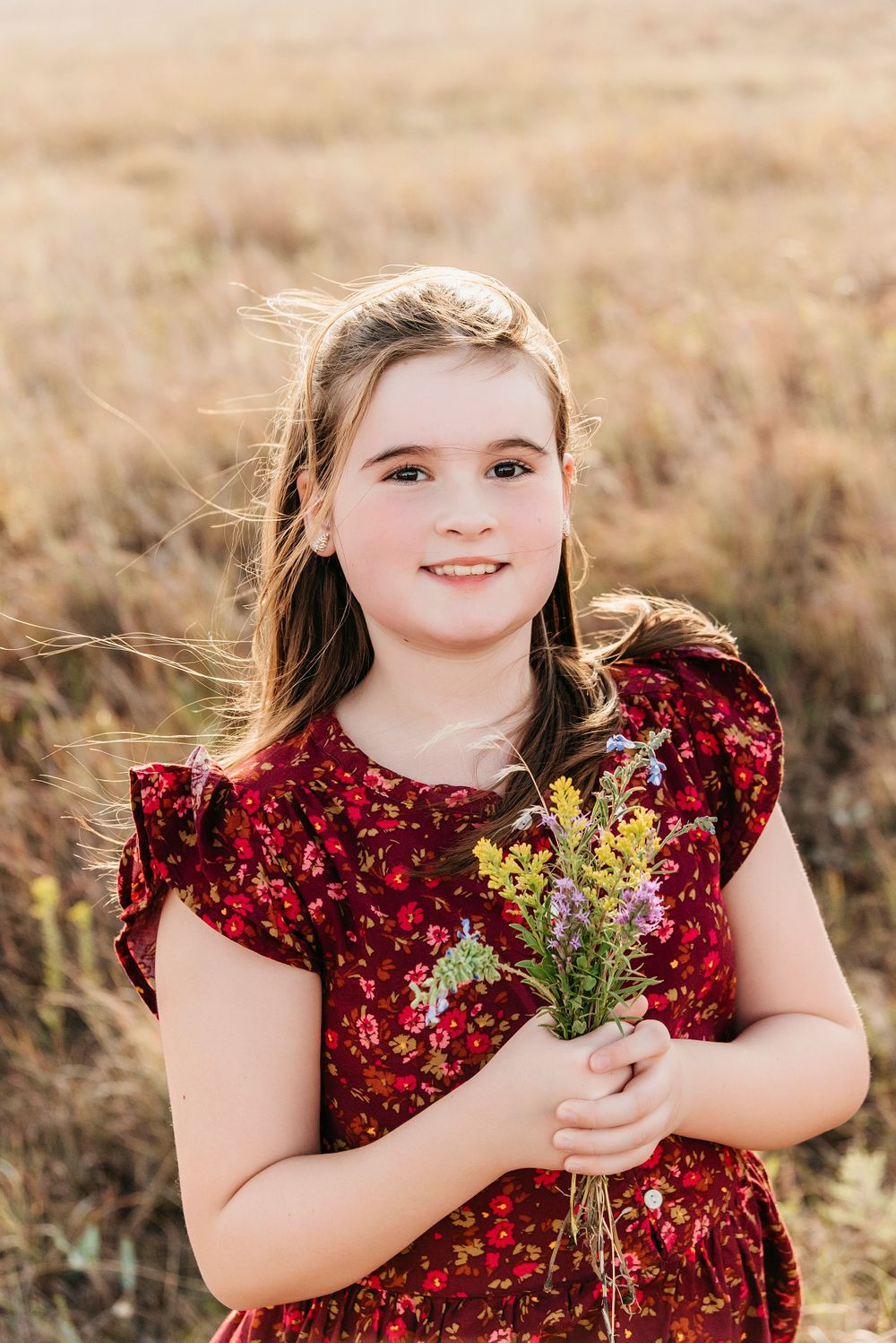 A child in a red sequin dress holds wildflowers while posing in a sunny field.