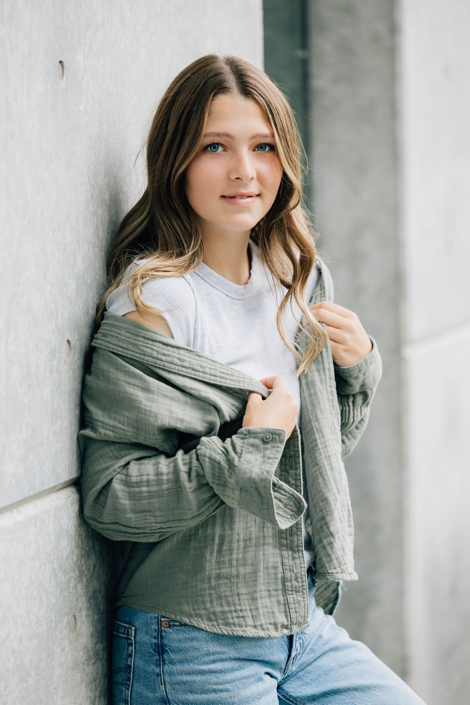 Teen girl in a green jacket and white tee leaning against a concrete wall.