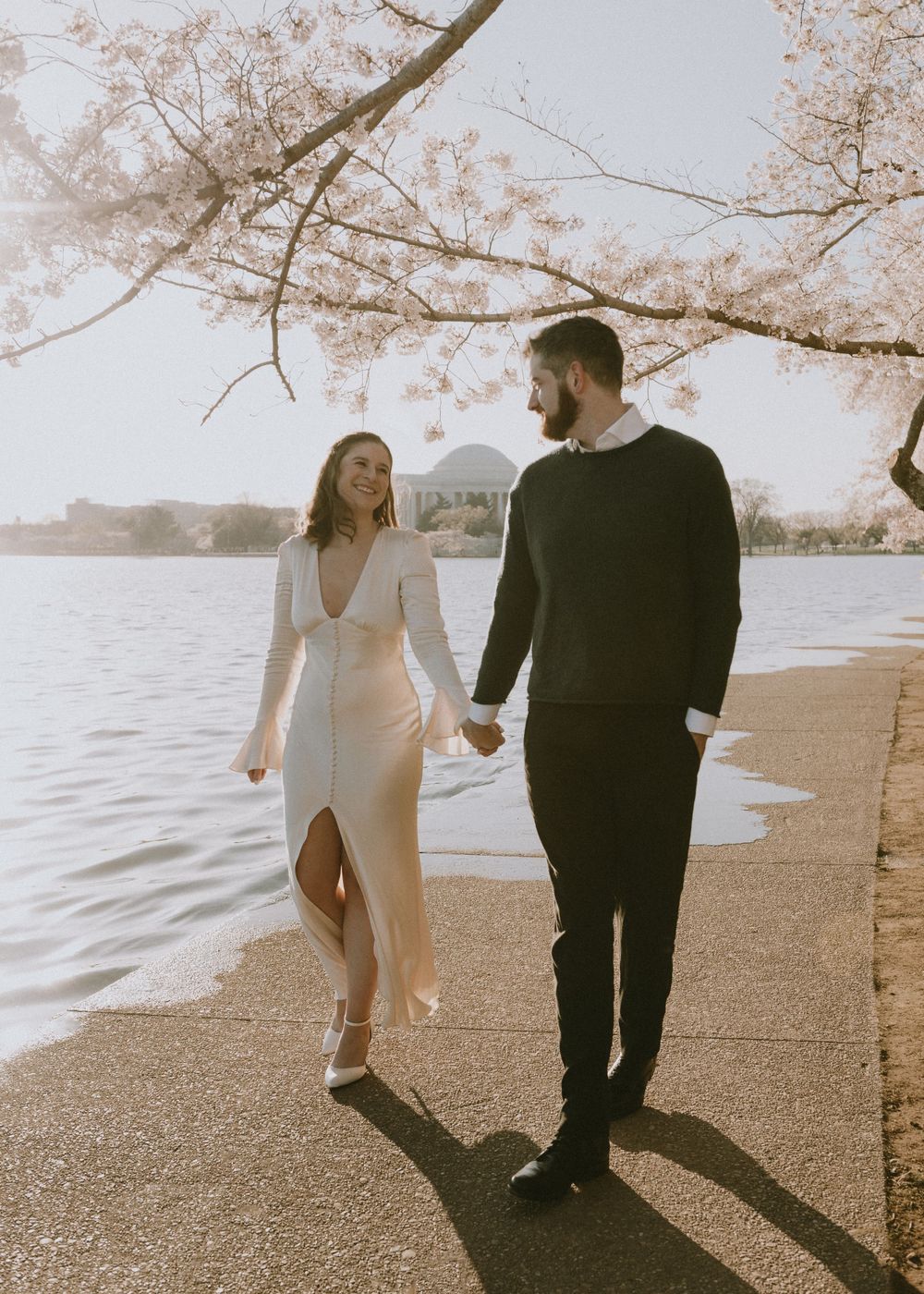 Couple walks hand in hand along waterfront path under blooming cherry blossoms.