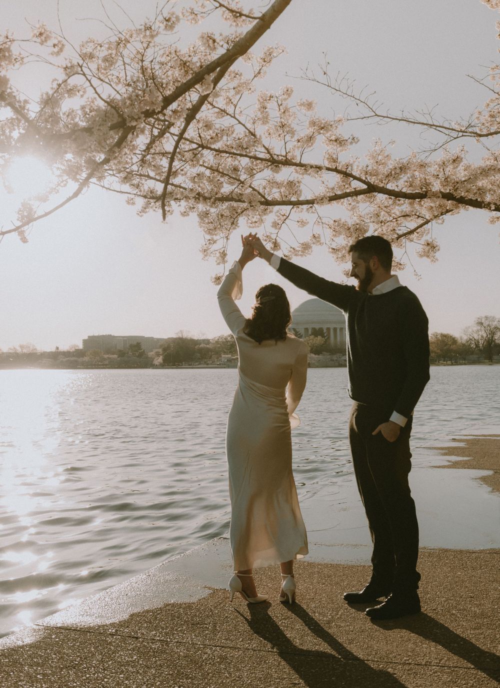 A couple dancing under blooming cherry blossom branches by the water at sunset.