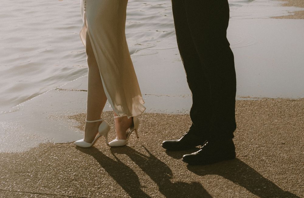 Close-up of formal shoes and dress shoes standing together on sandy ground near water.