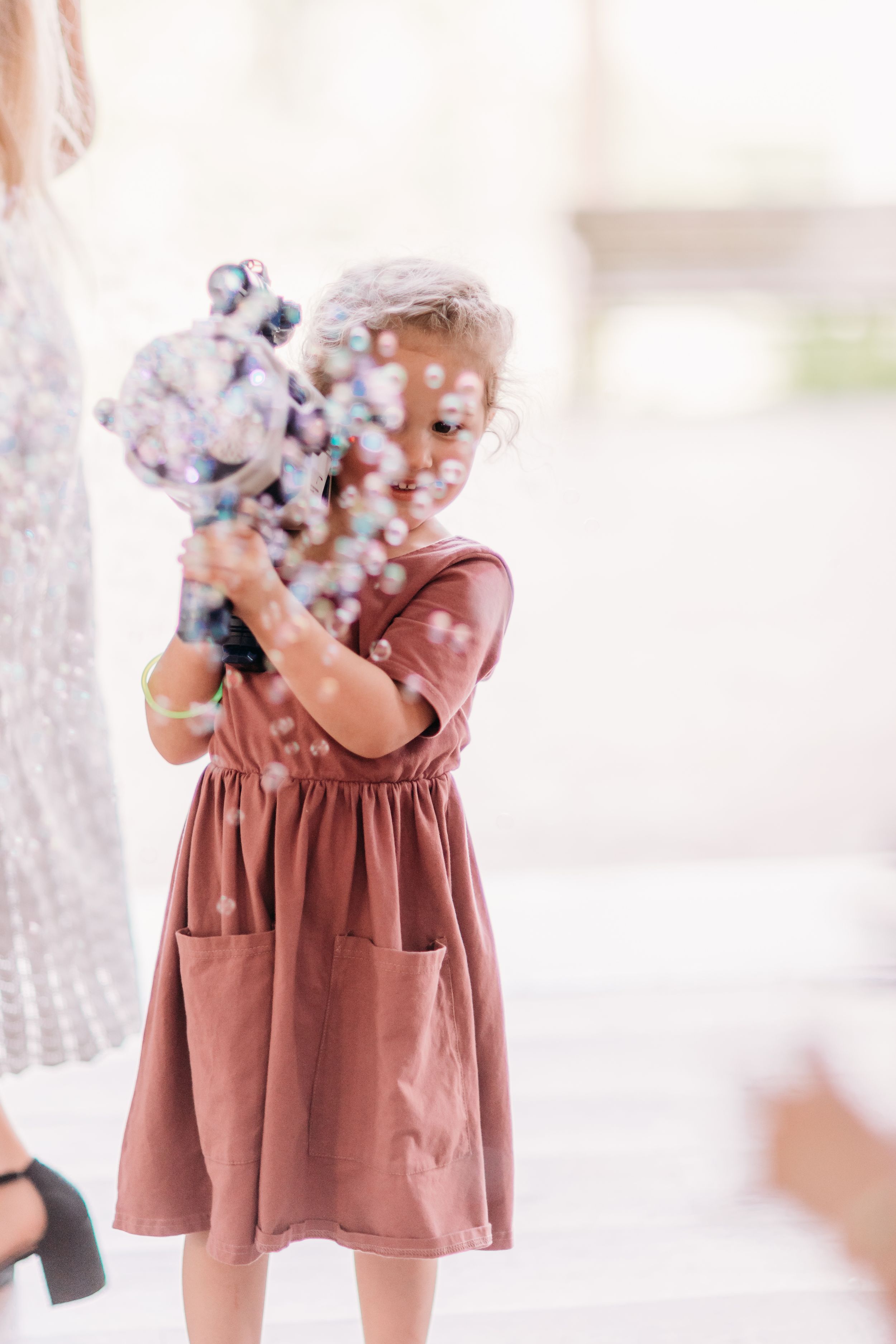 A young girl in a pink dress joyfully holds a bubble gun, surrounded by colorful bubbles outdoors.