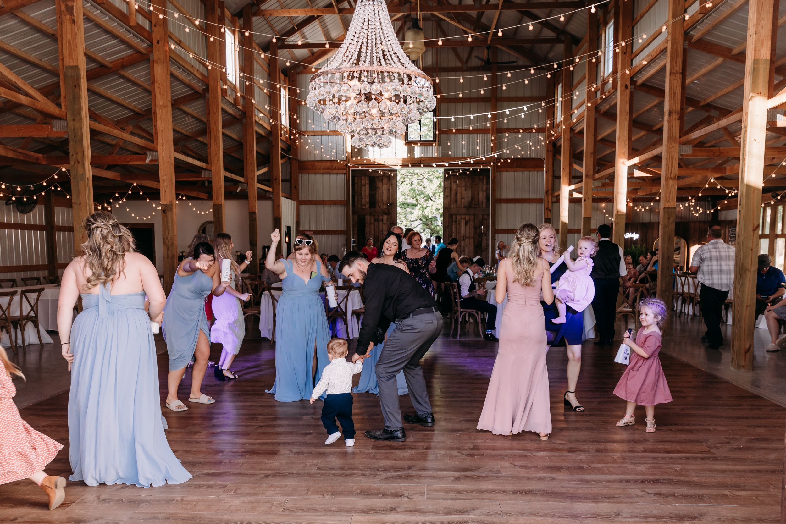 Guests dancing joyfully in a rustic barn wedding reception venue adorned with a crystal chandelier and string lights.