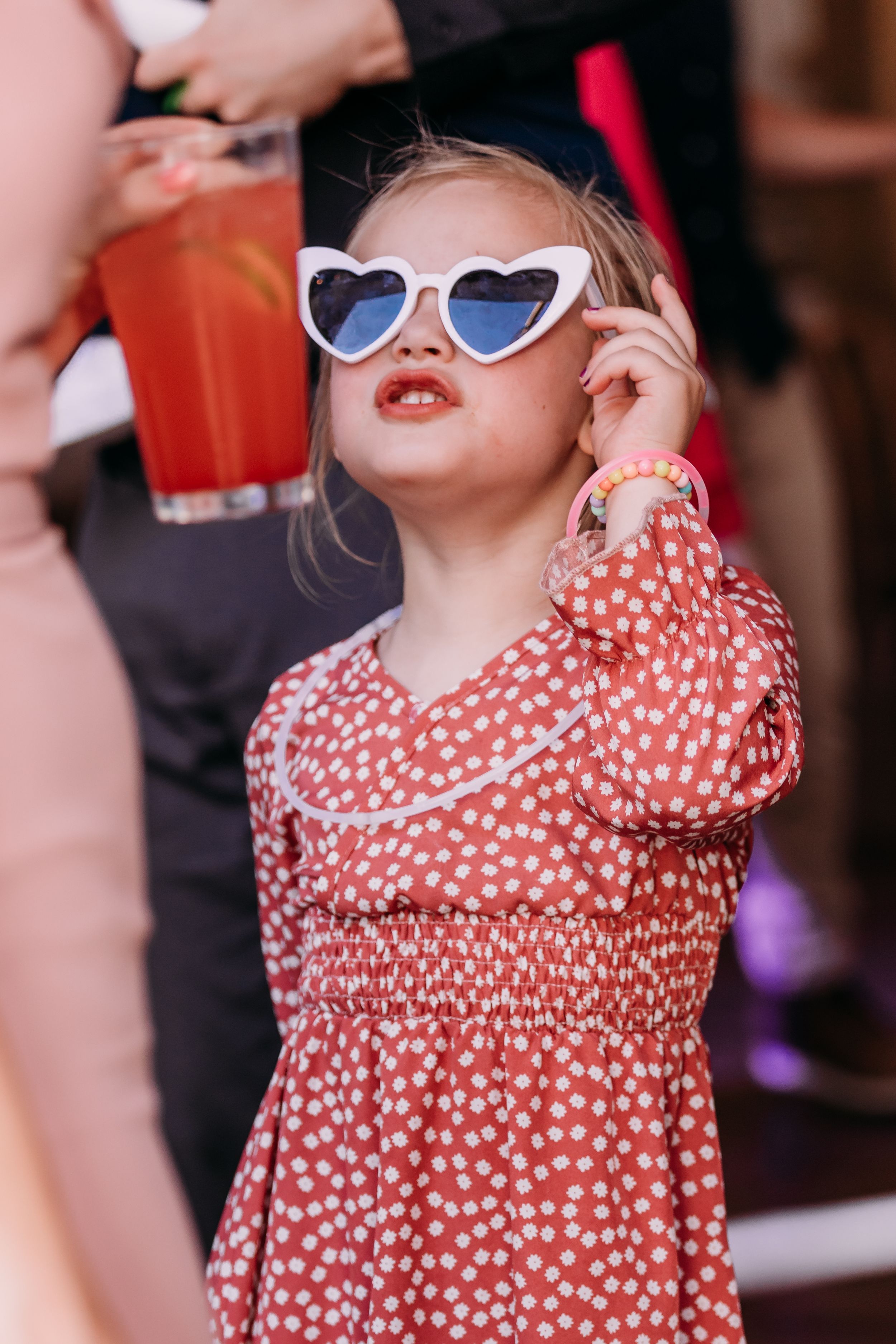 Cute little girl in red polka dot dress and white heart-shaped sunglasses looking up at a party.