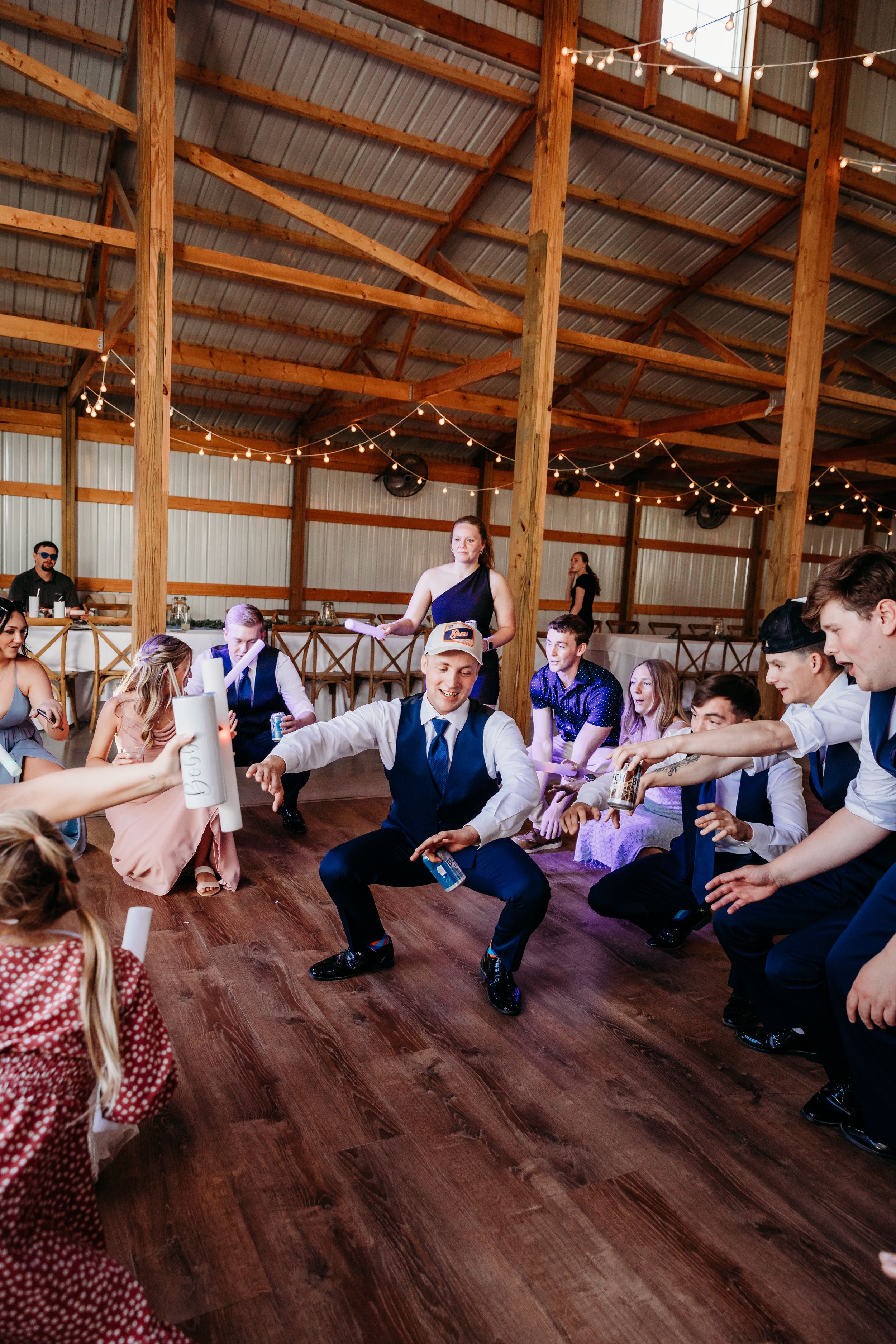 Wedding guests dancing in a rustic barn venue with string lights and wooden beams during a lively reception.