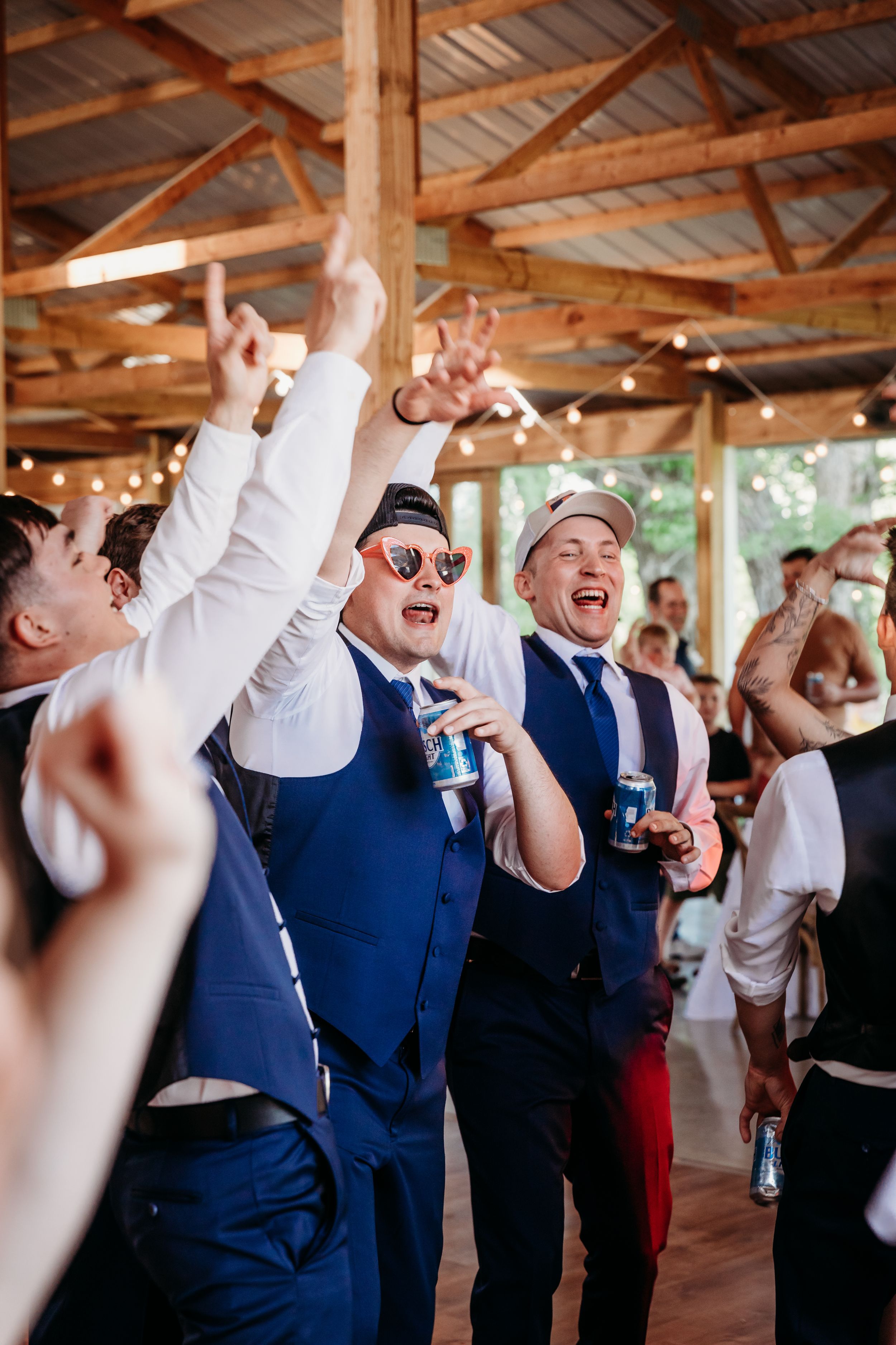 Men in blue vests celebrate at a rustic wedding reception, holding drinks and dancing under string lights.