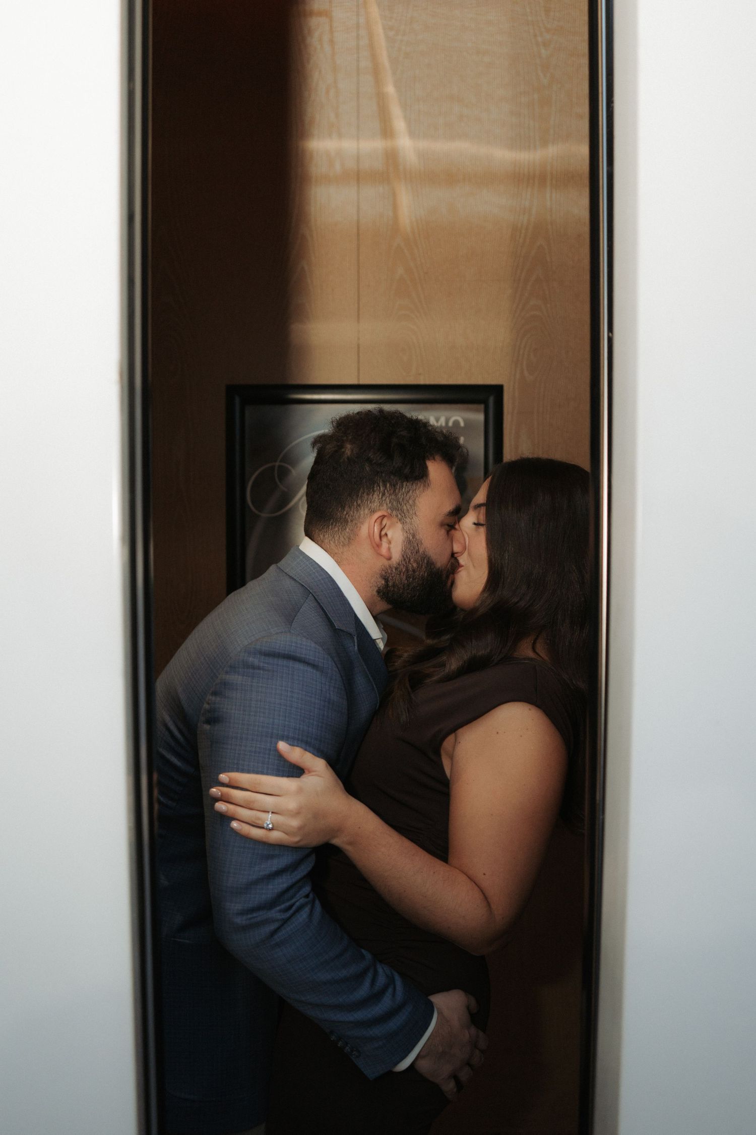 Couple sharing a romantic kiss in an elevator doorway, man in blue suit embracing woman in dark dress.