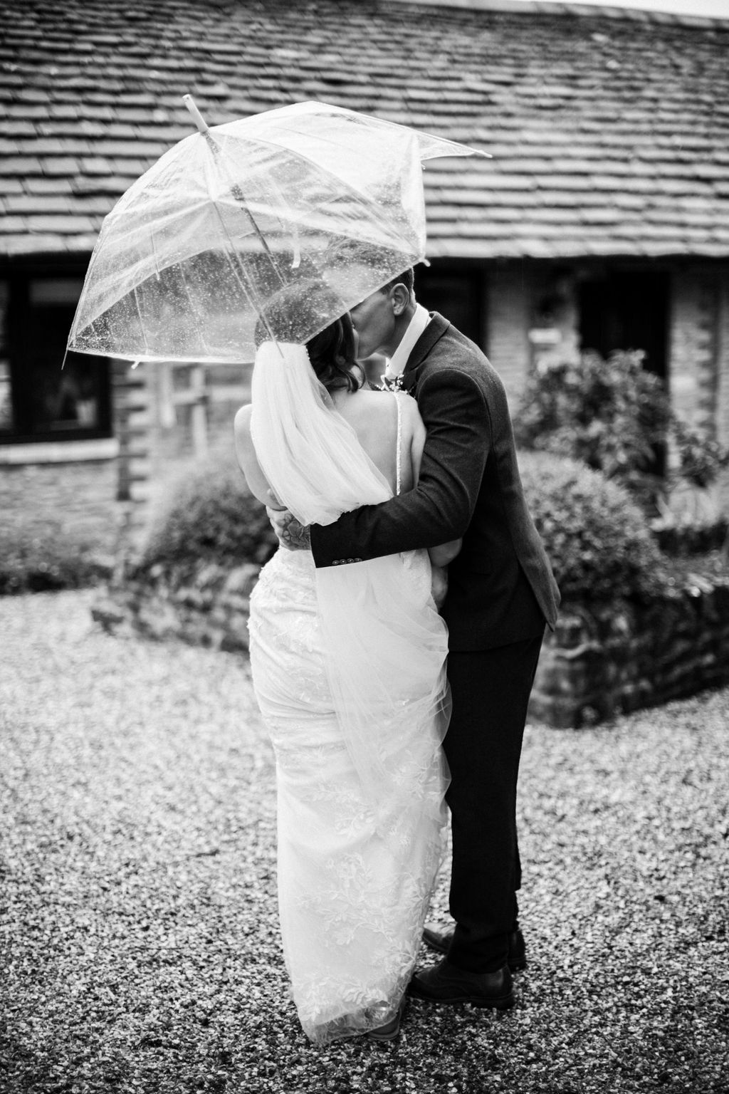 Black and white photo of bride and groom kissing under a clear umbrella on a rainy wedding day.