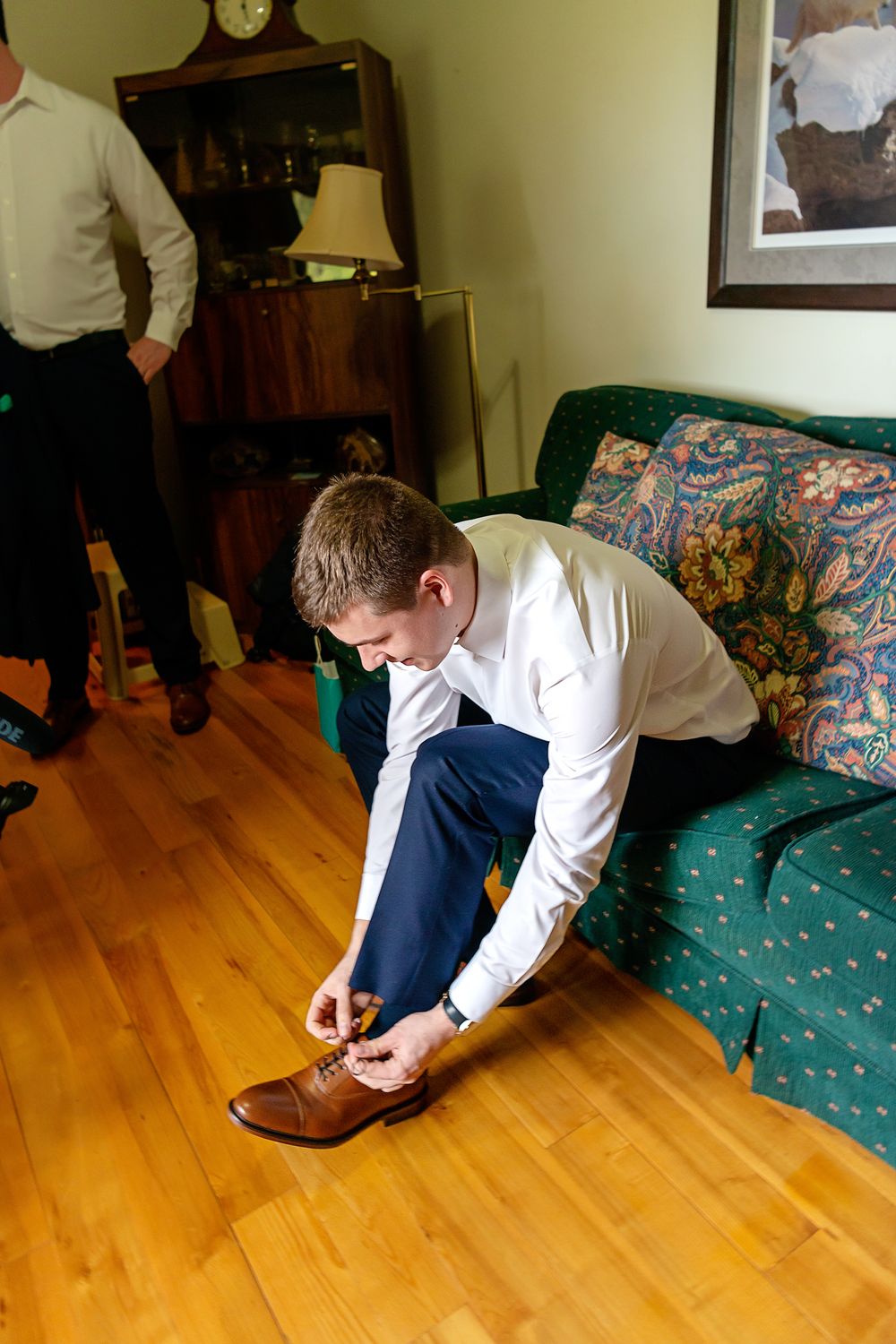 Groom in white shirt and blue pants tying brown dress shoes on hardwood floor before wedding ceremony.