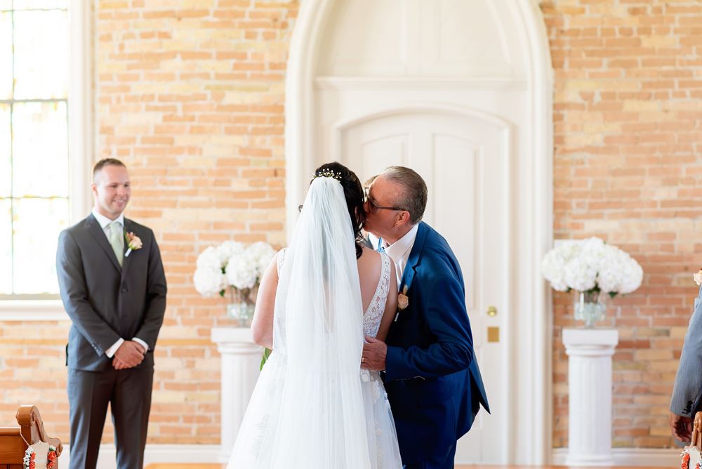 Bride and groom share first kiss at wedding altar in elegant chapel with brick walls and white floral arrangements.
